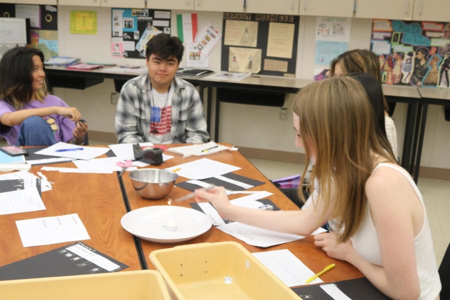 Fashion & Design students Desiree Dong and Zachary Smallridge work on papers and watch as Brooke Williams burns a piece of fabric over a candle.