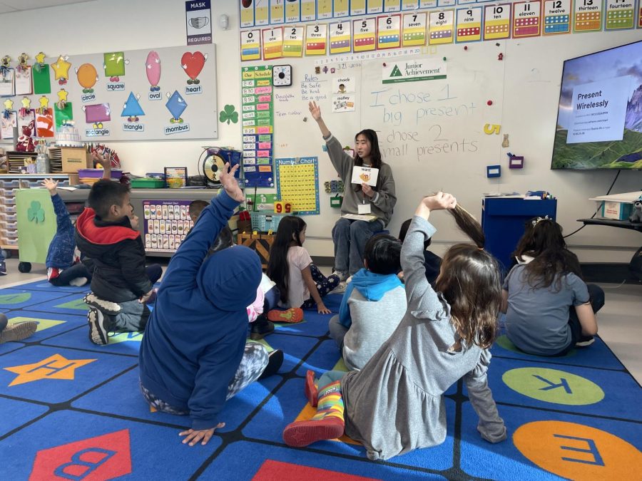 Michelle Obama Elementary School students sit around junior Hillary Kim as she teaches them different verbs. The children raise their hands to participate in this activity.