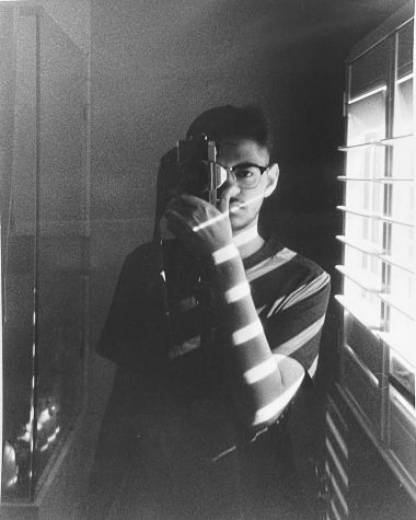 Junior Raza Ali takes a mirror self-portrait covering half his face with a camera.  He plays with the lighting by opening the blinds to bring contrast and shadows to his portrait.
