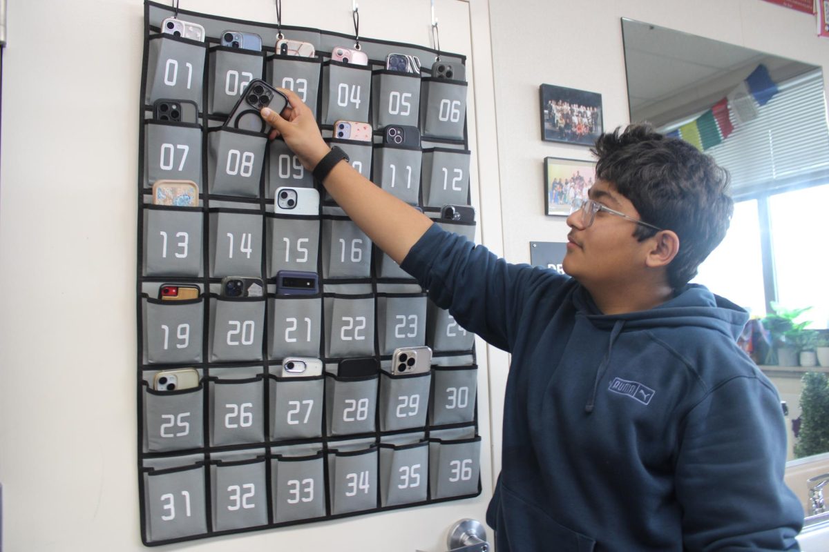 Freshman Hari Kotecha reluctantly puts his phone into Laura Woods’ class phone caddy. Starting this semester, students are required by the school to put their phones in these caddies. Last semester, students could store them in their backpacks.